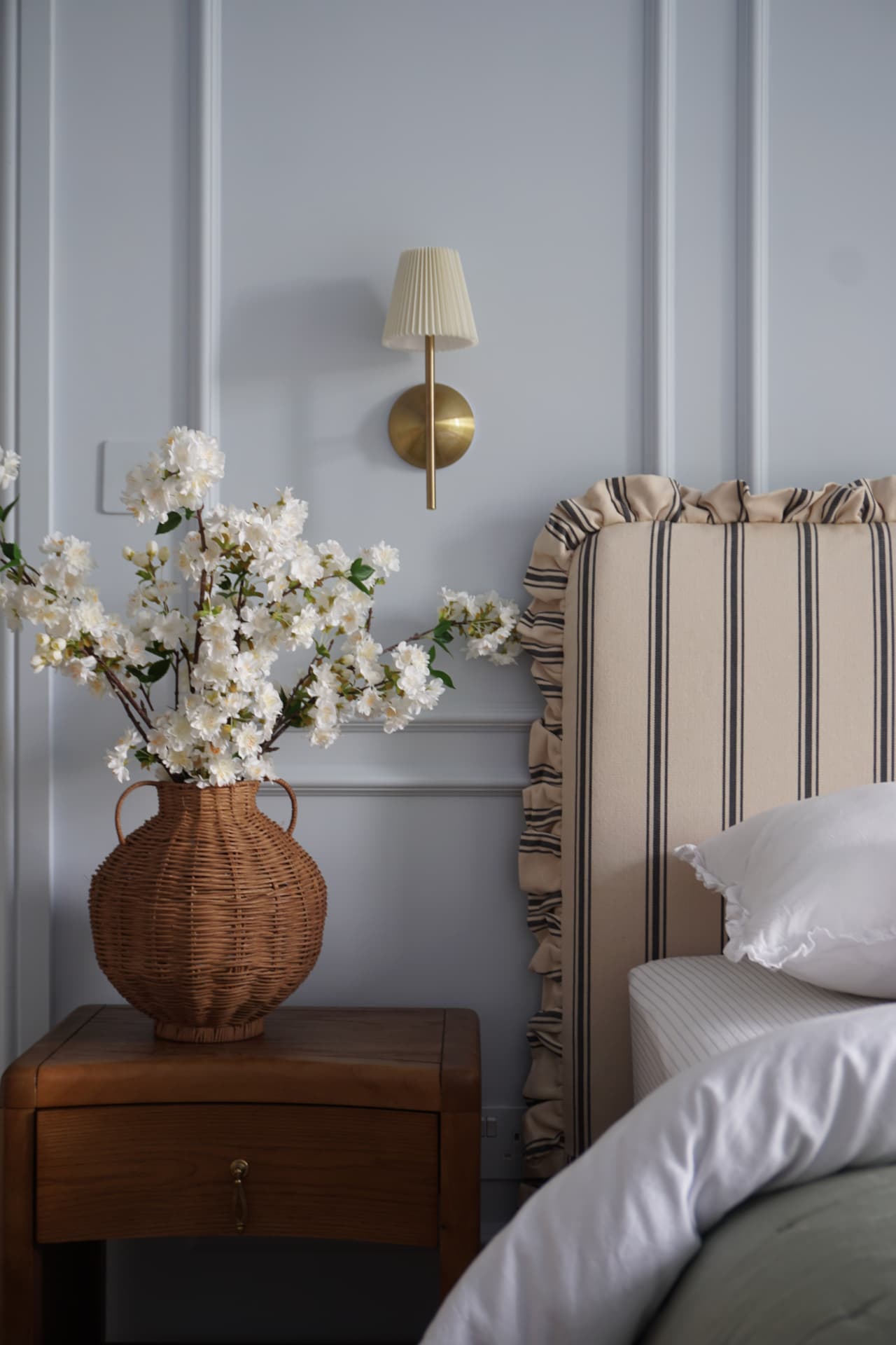 Rattan vase of white blossom on a wooden bedside table against a blue panelled wall